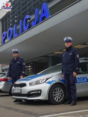 Policjanci stoją przy radiowozie na tle budynku komendy