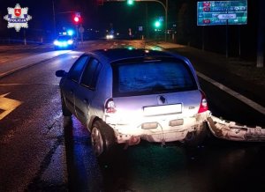 Uszkodzony samochód marki renault stojący przed sygnalizacją świetlną. W oddali radiowóz policyjny