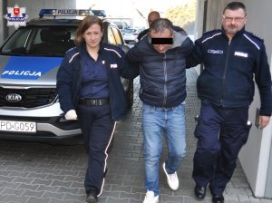 Mężczyzna w czarnej kurtce prowadzony przez policjantów. W tle radiowóz