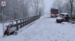 Uszkodzony samochód stoi na jezdni. Za nim wóz Straży Pożarnej z włączonymi światłami błyskowymi.