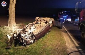 Uszkodzony samochód leży na dachu. W tle policjant