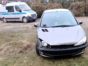 Uszkodzony peugeot stoi na trawiastym podłożu. Za nim oznakowany radiowóz policyjny