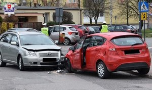 Uszkodzone auta stoją na łuku drogi. W tle policjanci.