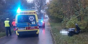 Miejsce zdarzenia. Na trawiastym podłożu stoi motorower. Na jezdni znajduję się karetka pogotowia i radiowóz policyjny