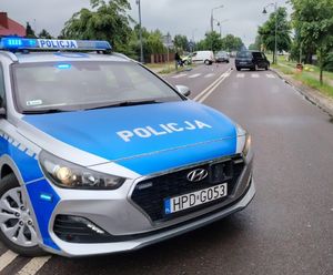 Oznakowany radiowóz stoi na jezdni. Policjanci zabezpieczają miejsce wypadku drogowego
