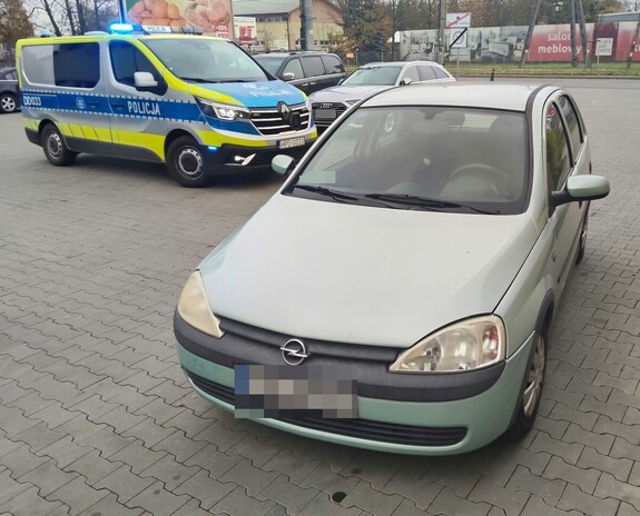 Samochód marki Opel Corsa stoi na parkingu, a za nim oznakowany radiowóz